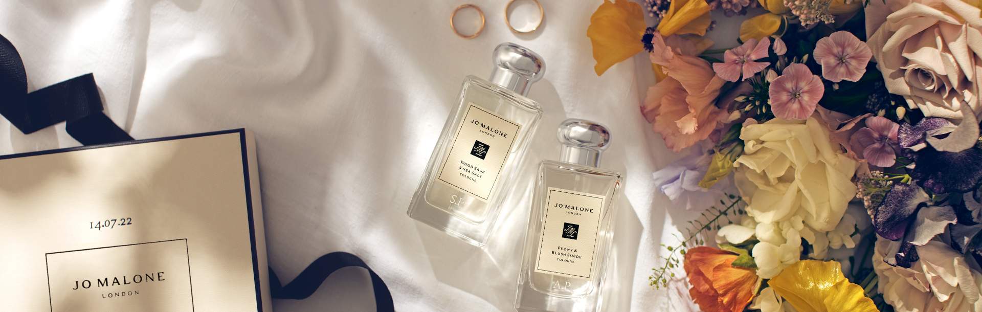 Image of two engraved jo malone colognes next to gold wedding rings & colourful flowers